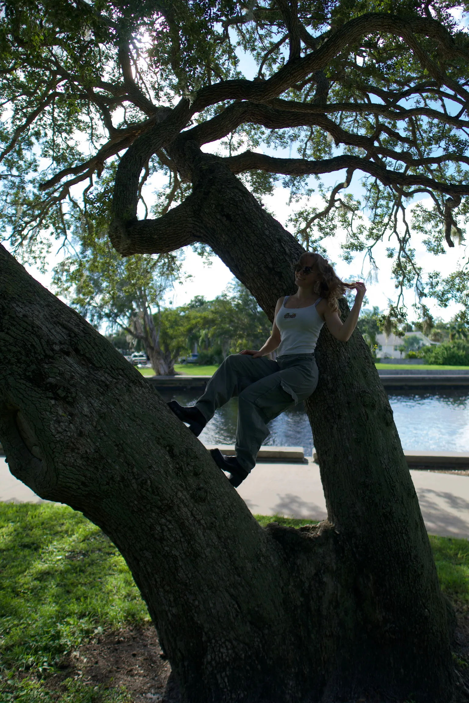 A woman leaning against the trunk of a leaning live oak beside a waterway, seen from a distance
