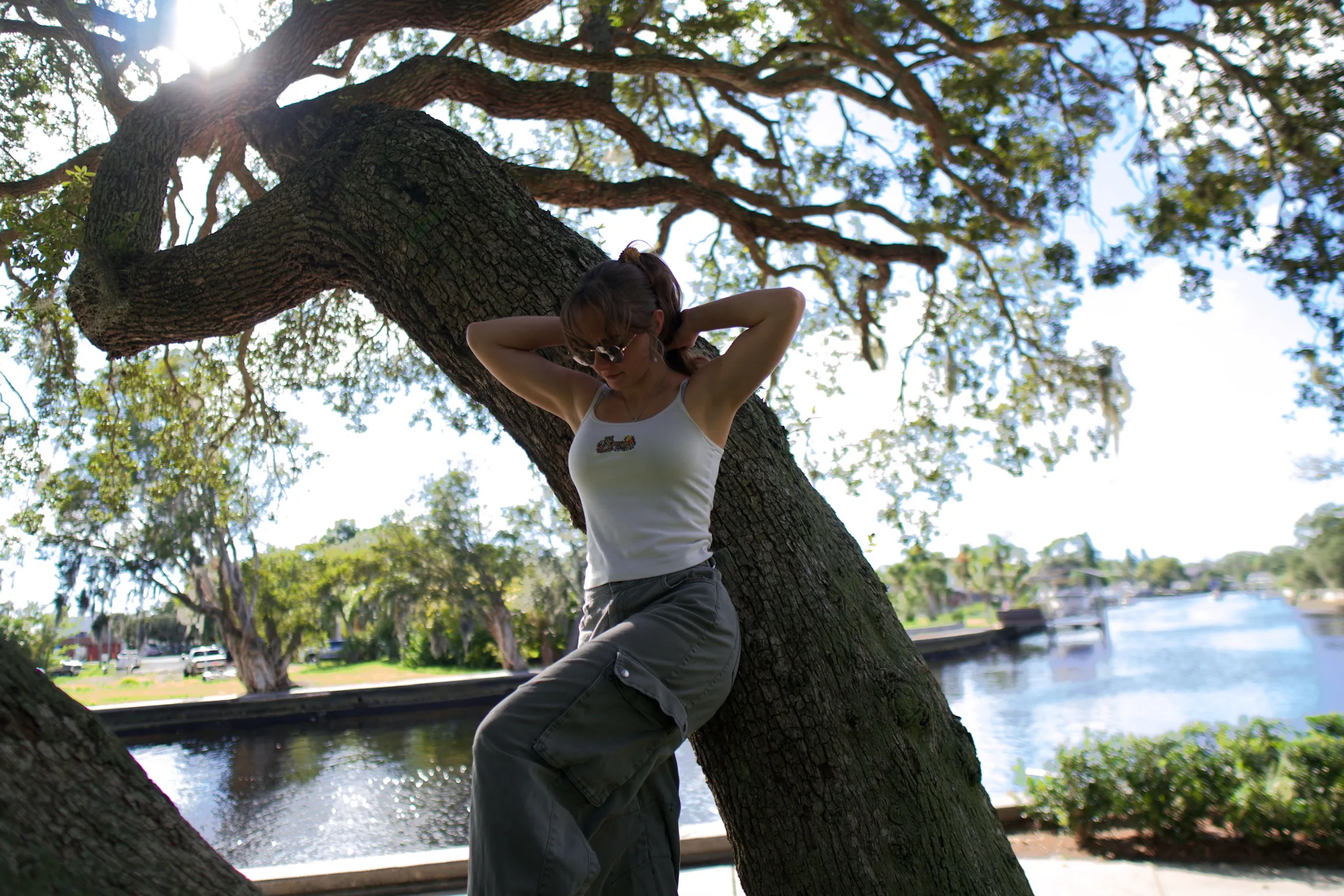 A woman sitting on a low oak branch with her hands behind her head, sun through the canopy