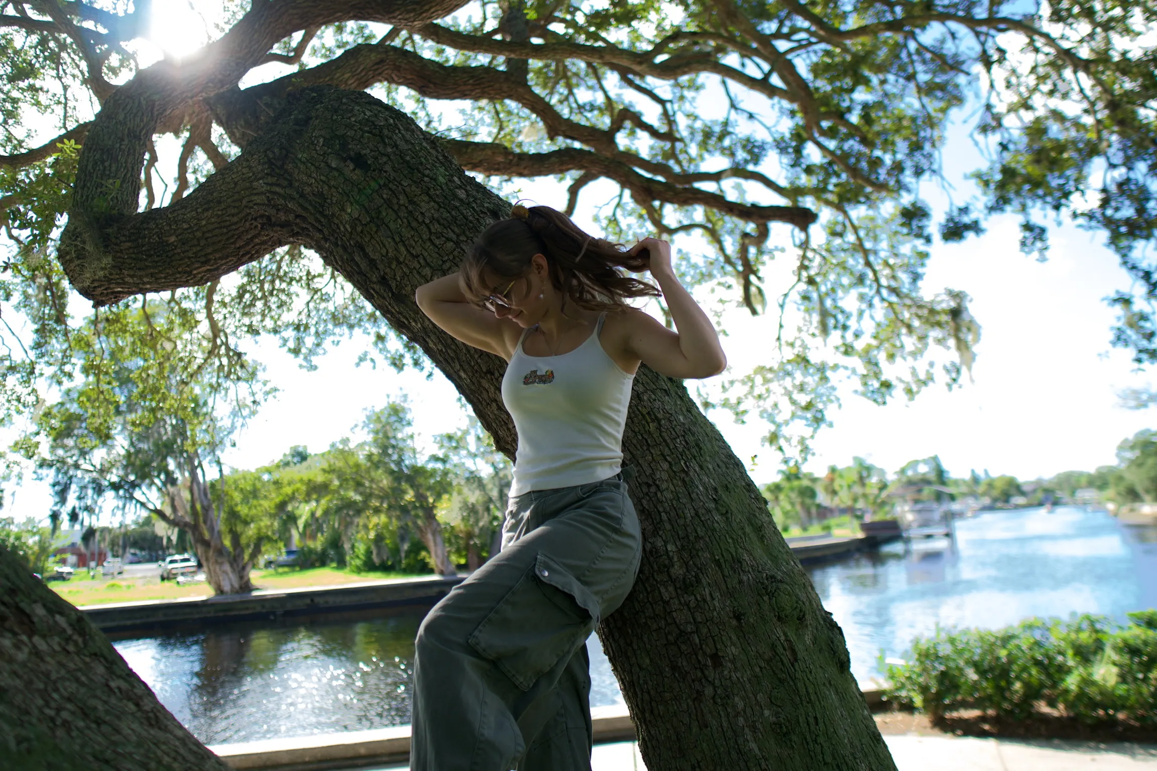 A woman sitting on a low oak branch, hands behind her head, water and boats behind