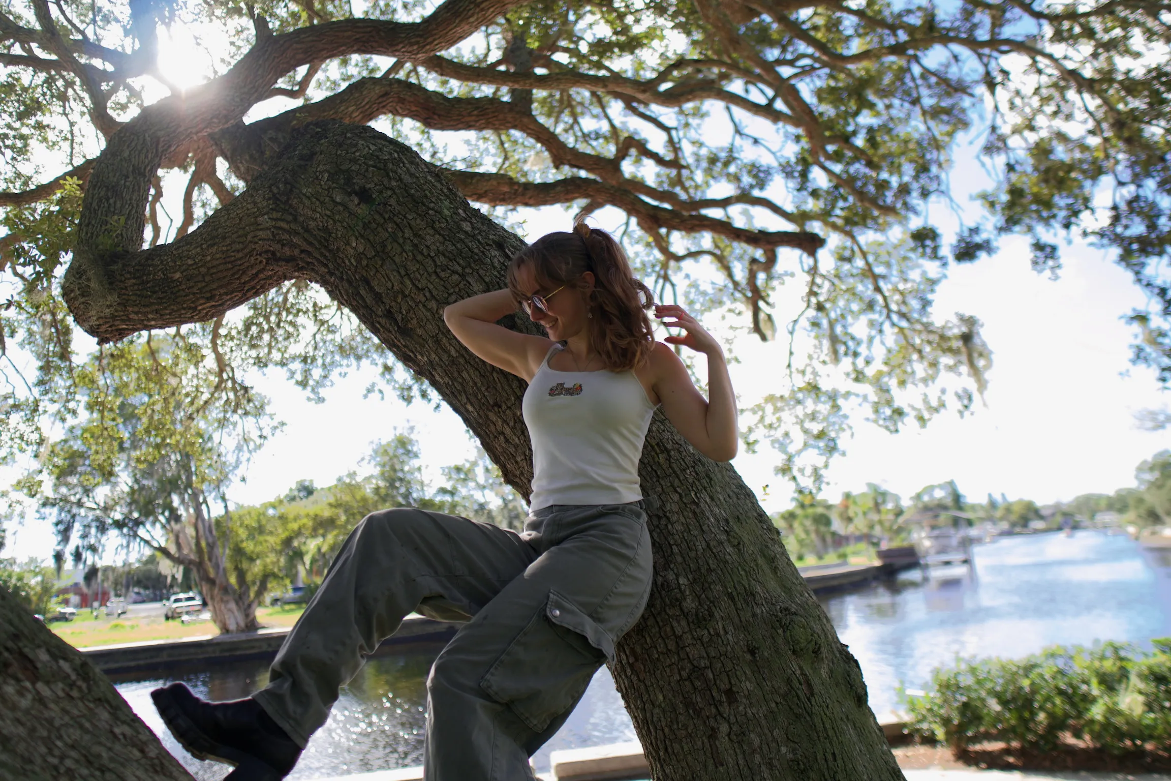 A woman seated on a leaning oak trunk with one hand in her hair, marina in the background