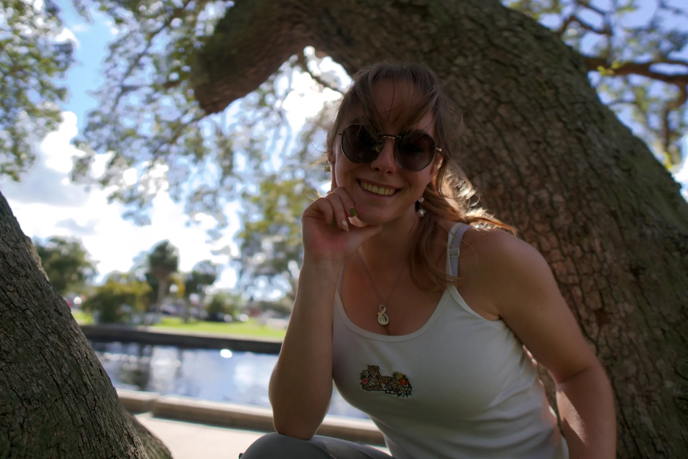 Close portrait of a woman in round sunglasses smiling under an oak, clouds behind
