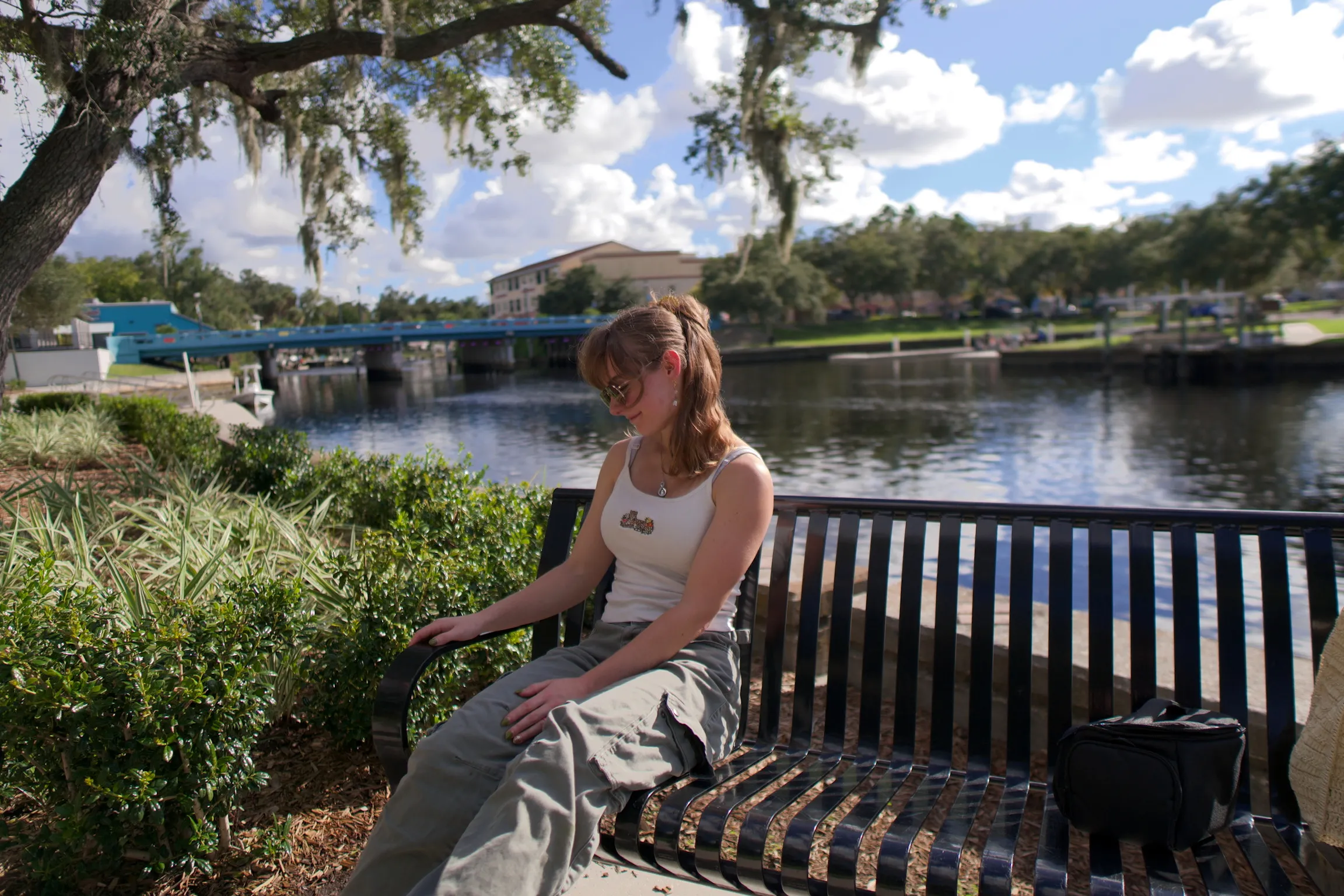 A woman on a bench by the water, looking down, blue sky and clouds