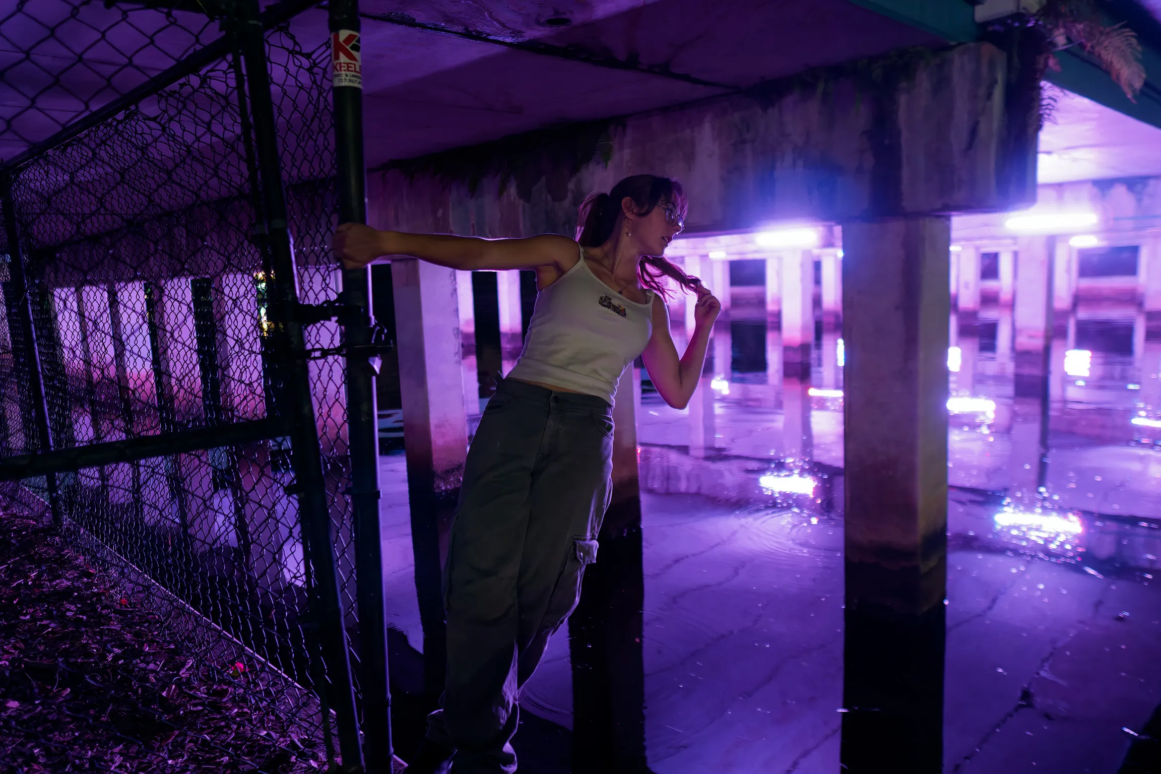A woman at night with her arms out along a chain-link fence, purple light under a pier