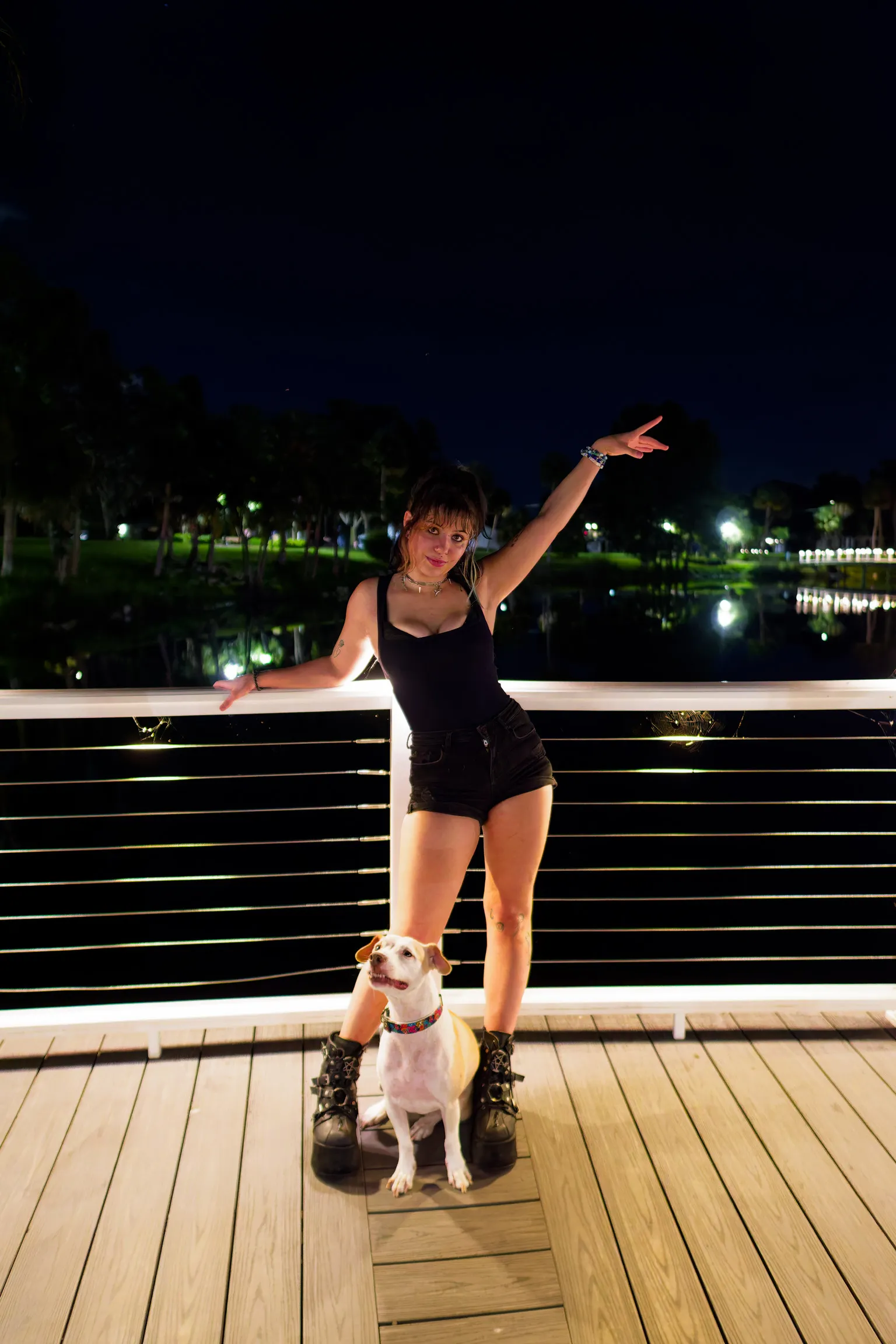 A woman at a boardwalk railing at night with one arm raised, small white dog at her feet